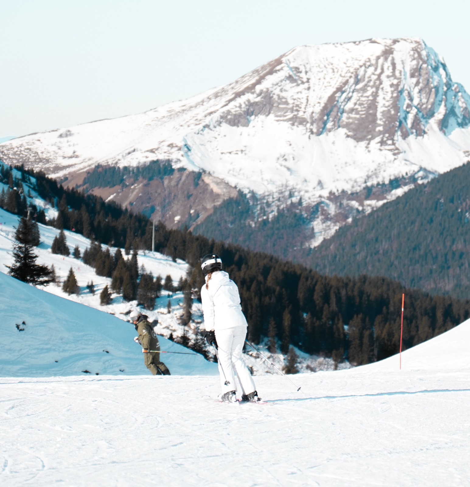 A Peak instructor and client pause on piste, an Alpine peak rising behind them.